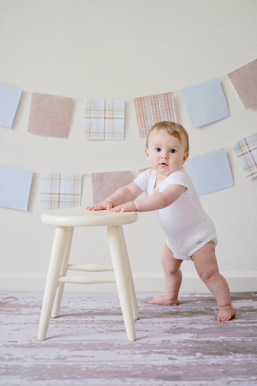 baby holding white wooden stool|unrecognizable woman arranging baby clothes at table