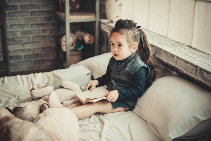 toddler girl wearing long sleeved top reading book while sitting on bed